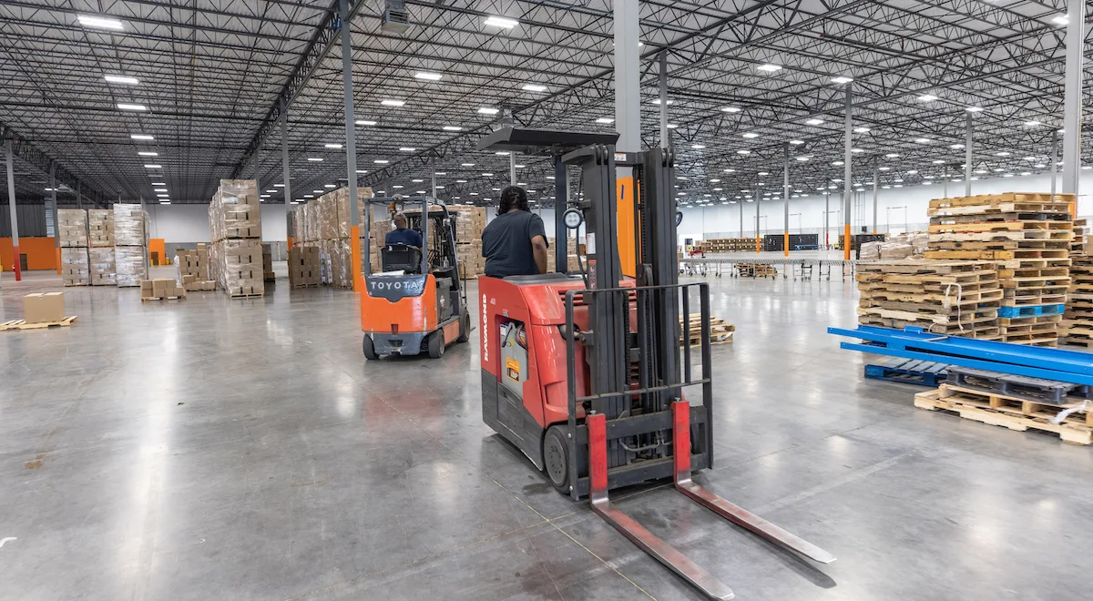 Two forklifts with operators in a warehouse with boxes and pallets.